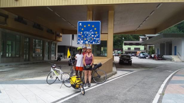 Drei Radfahrer posieren unter einem Schild, das die italienische Grenze markiert.