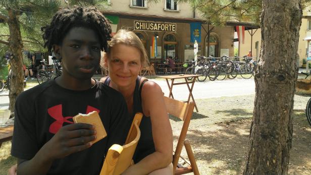 Ein junger Mann isst ein Eis, während eine Frau neben ihm sitzt; im Hintergrund Fahrräder und das Schild „Chiusaforte“.