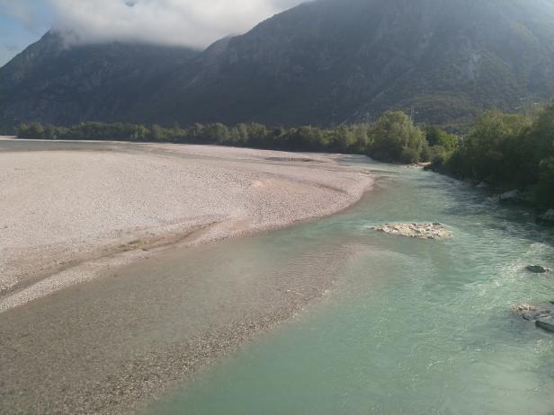 Ein Fluss mit türkisfarbenem Wasser fließt durch eine Kiesbank vor einer Bergkulisse.