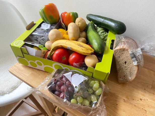 Ein Korb mit frischem Obst und Gemüse, darunter Paprika, Kartoffeln, Zucchini, Bananen, Tomaten, Trauben und Brot.