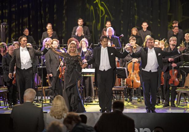 Eine Sängerin und zwei Sänger stehen mit einem Orchester auf der Bühne.