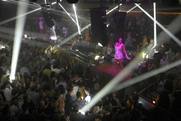 Eine Frau in einem glitzernden Kleid steht auf einer Bühne in einem Nachtclub, umgeben von einer tanzenden Menschenmenge.