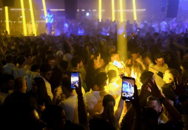 Eine große Menschenmenge feiert in einem Club mit gelben Lichtern und erhobenen Handys.