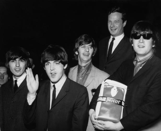 Die Beatles posieren mit einer Tasche mit der Aufschrift „John“ für ein Schwarzweißfoto.