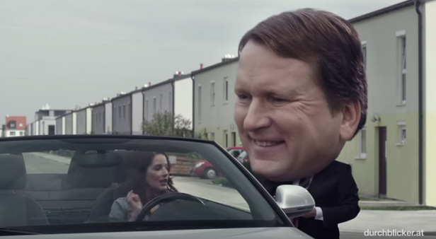 Eine Frau fährt ein Auto, während ein Mann mit übergroßem Kopf am Fenster steht.