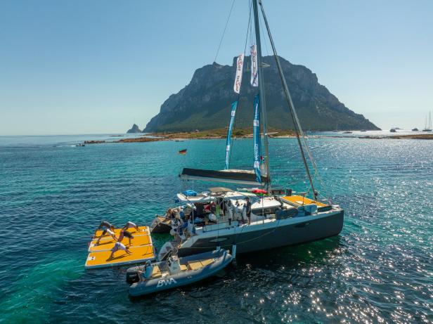 Ein Katamaran ankert vor einer Insel, während eine Gruppe Yoga auf einer Plattform im Wasser praktiziert.