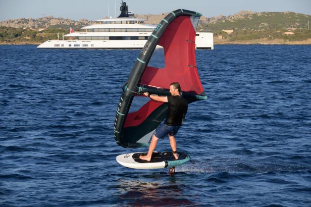 Ein Mann wingfoilt auf dem Meer, im Hintergrund eine große Yacht.