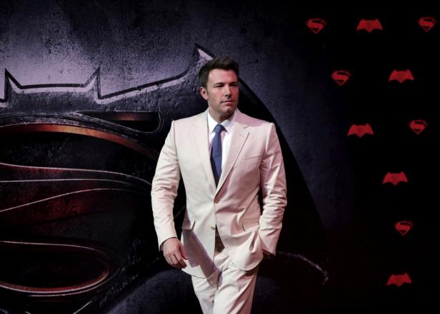 Ben Affleck posiert vor einem Hintergrund mit Batman- und Superman-Logos.