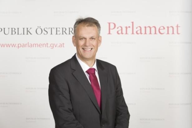 Ein lächelnder Mann im Anzug vor dem Hintergrund des österreichischen Parlaments.