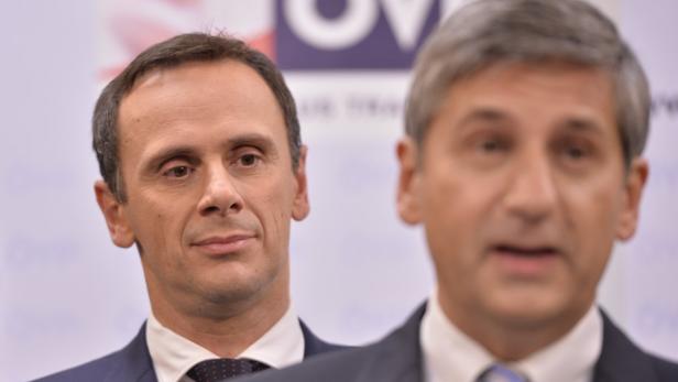 Zwei Männer in Anzügen stehen vor einer Wand mit einem Logo.