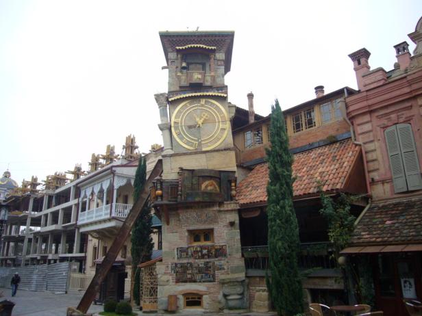 Ein schiefer Uhrturm mit goldenem Zifferblatt steht vor einer Reihe von Gebäuden in Tiflis, Georgien.