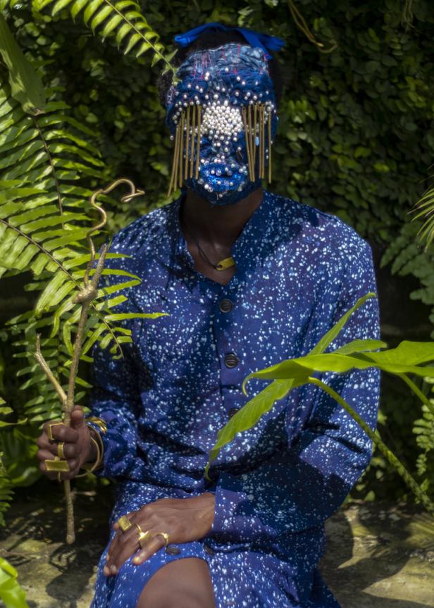 Eine Person mit einer verzierten Maske und blauem Hemd posiert inmitten von üppigem Grün.