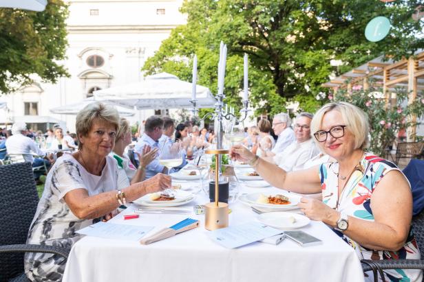 Zwei Frauen sitzen an einem Tisch bei einer Veranstaltung im Freien und halten Weingläser hoch.