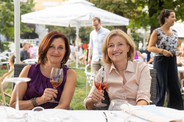 Zwei Frauen sitzen an einem Tisch im Freien und halten ihre Getränke.