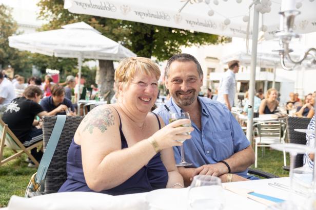 Ein lächelndes Paar sitzt an einem Tisch im Freien, die Frau hält ein Weinglas.