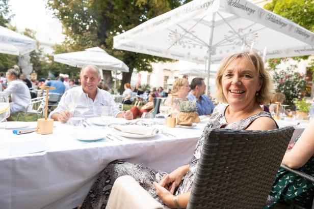 Eine lächelnde Frau sitzt an einem Tisch im Freien unter einem Sonnenschirm.