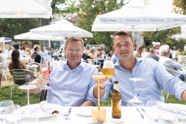 Zwei Männer sitzen an einem Tisch im Freien und halten Getränke in der Hand.