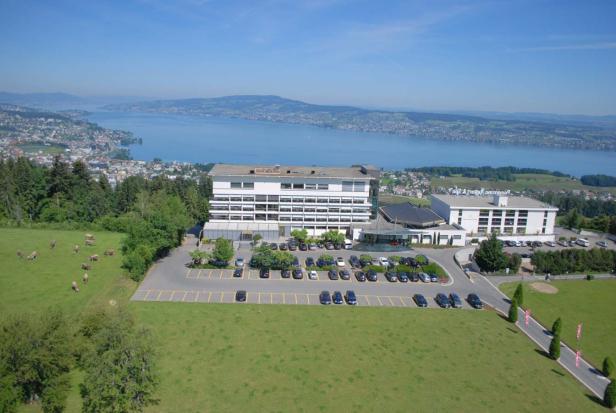 Ein Hotel mit Parkplatz und Blick auf einen See und bewaldete Hügel.