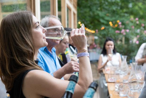 Eine Frau trinkt ein Glas Weißwein im Freien, mit anderen Personen im Hintergrund.