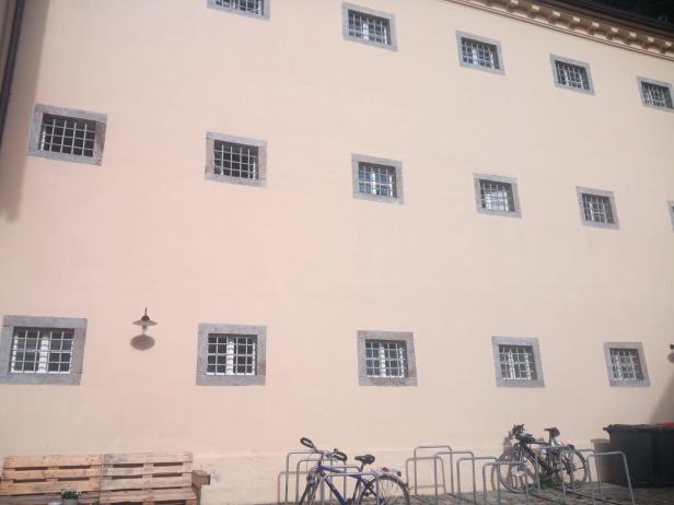 Eine Fassade mit vergitterten Fenstern, Fahrrädern und Paletten im Vordergrund.