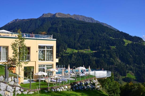 Ein Hotel mit Pool und Liegestühlen vor einer bewaldeten Bergkulisse.