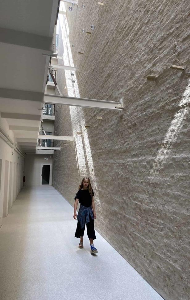Ein junges Mädchen geht in einem langen Flur mit einer hohen Ziegelwand entlang.