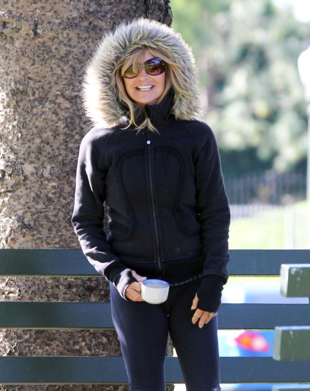 Eine Frau mit Sonnenbrille und Kapuzenjacke hält eine Tasse in der Hand.