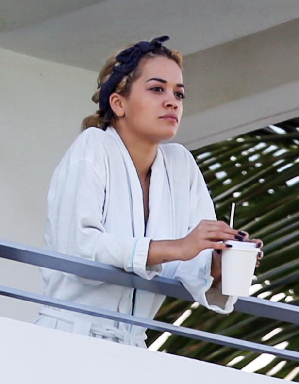 Rita Ora in einem weißen Bademantel auf einem Balkon mit einem Getränk in der Hand.