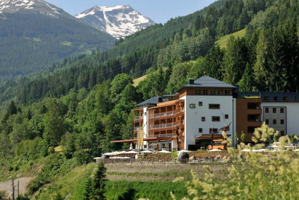 Ein Hotel liegt vor einer bewaldeten Berglandschaft mit schneebedeckten Gipfeln.