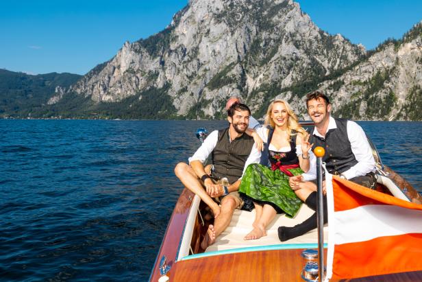 Drei Personen in Tracht sitzen in einem Boot auf einem See vor einer Bergkulisse.