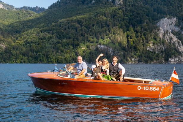 Vier Personen fahren mit einem Holzboot auf einem See vor einer bewaldeten Bergkulisse.