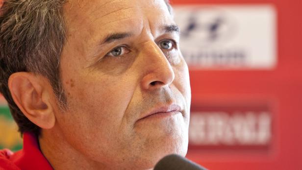 Ein Mann mittleren Alters mit ernstem Gesichtsausdruck vor rotem Hintergrund.