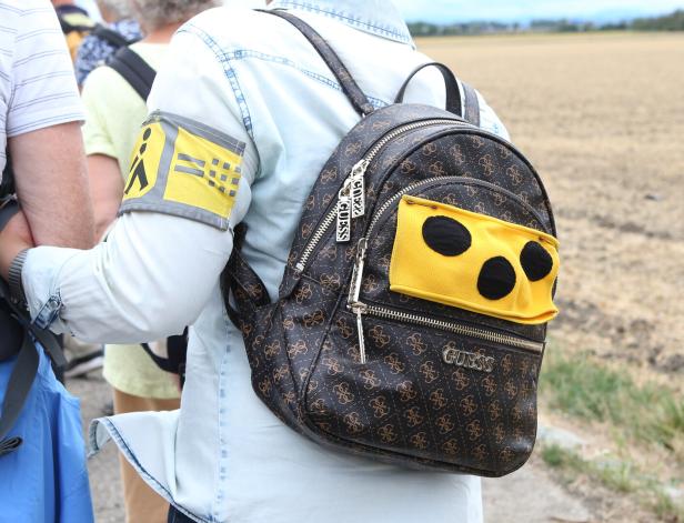 Person mit Rucksack und gelbem Armband mit Symbolen, im Freien auf einem Feldweg unterwegs.