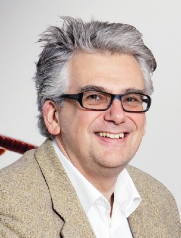 Ein lächelnder Mann mit Brille und grauem Haar trägt ein beiges Jackett.