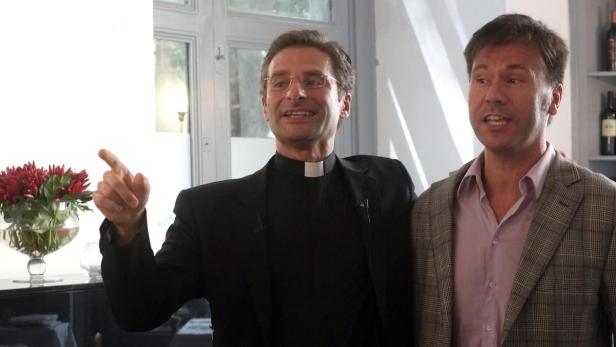 Ein Priester gestikuliert neben einem Mann mit kariertem Sakko.