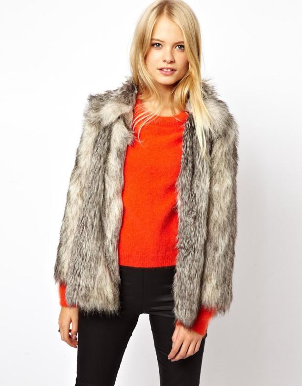 Eine blonde Frau trägt einen orangefarbenen Pullover und eine graue Pelzjacke.