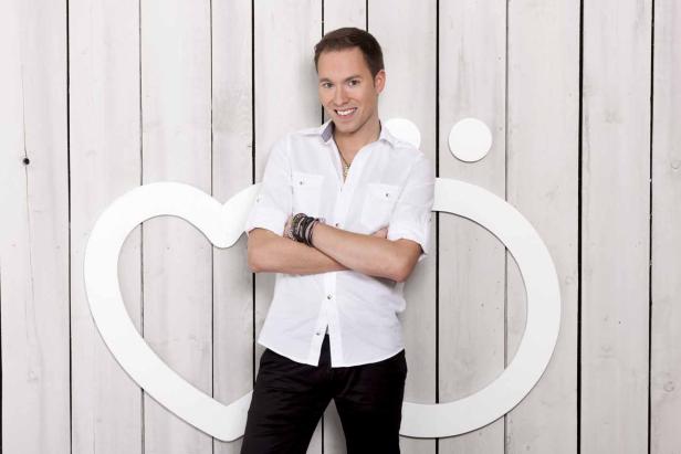 Ein Mann mit verschränkten Armen vor einem weißen Herz- und Kreissymbol auf einer Holzwand.