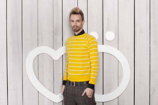 Ein Mann mit gelb-weiß gestreiftem Pullover posiert vor einer Wand mit Herz- und Kreismotiven.