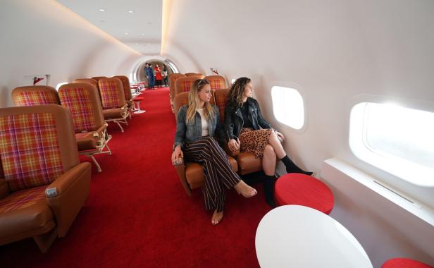 Zwei Frauen sitzen in einem Flugzeuginterieur mit roten Teppichen und blicken aus dem Fenster.