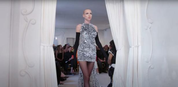 Ein Model präsentiert ein silbernes, paillettenbesetztes Minikleid mit Schleppe auf dem Laufsteg.