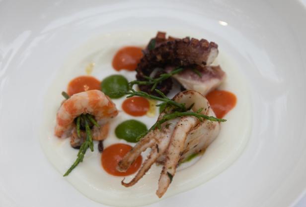 Ein Teller mit gegrilltem Tintenfisch, Garnele und Oktopus auf einem Bett aus Sauce, garniert mit Queller.