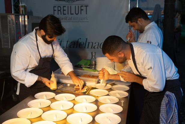 Drei Köche bereiten in einer Freiluftküche Teller mit Speisen zu.