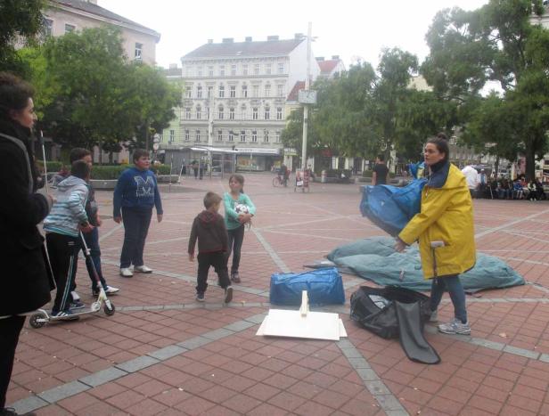 Eine Frau in einem gelben Regenmantel hantiert mit einer blauen Plane auf einem Platz.