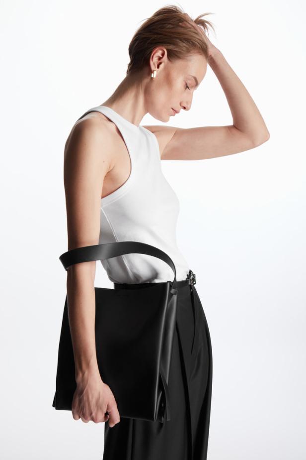 Eine Frau mit weißem Top und schwarzer Hose hält eine schwarze Handtasche.