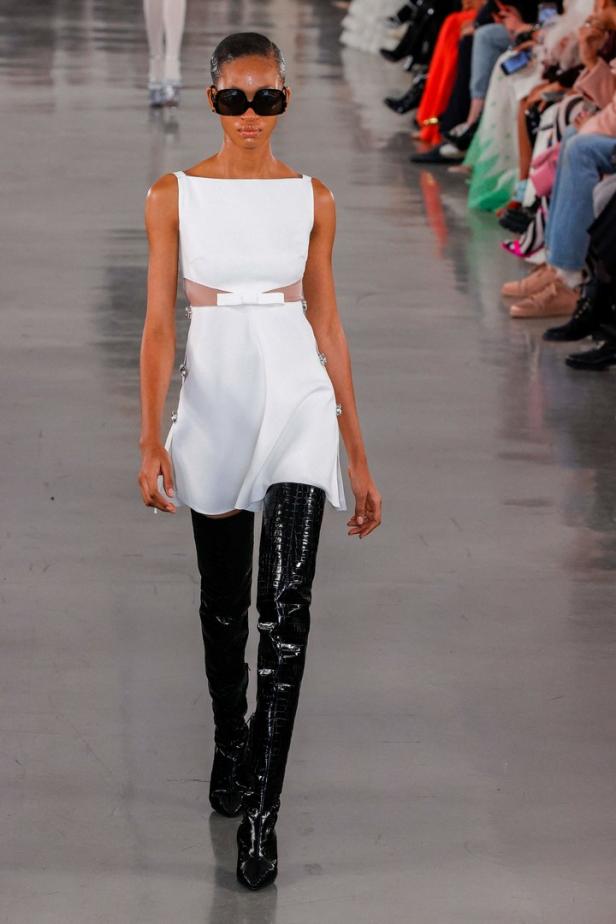 Ein Model mit Sonnenbrille präsentiert ein weißes Minikleid und schwarze Overknee-Stiefel auf dem Laufsteg.