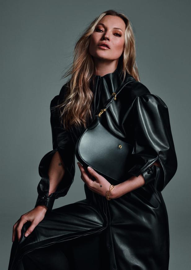 Kate Moss präsentiert eine schwarze Aigner-Handtasche und ein goldenes Armband.