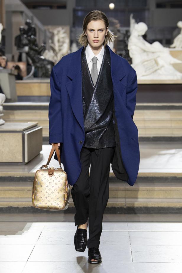 Ein Model in einem blauen Mantel, einem glitzernden Oberteil und einer Louis Vuitton Tasche auf einem Laufsteg.