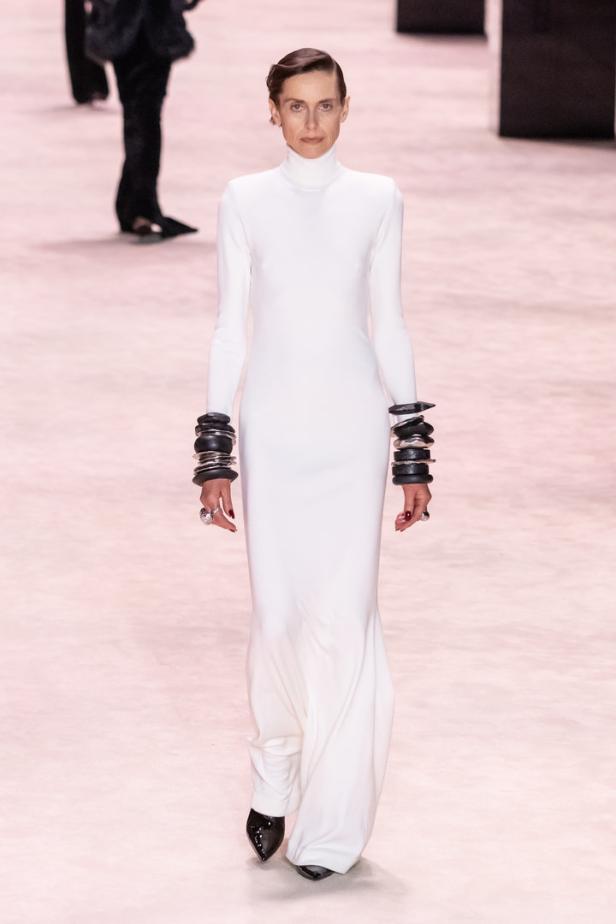 Eine Frau in einem weißen Kleid mit Rollkragen und auffälligen Armreifen auf dem Laufsteg.