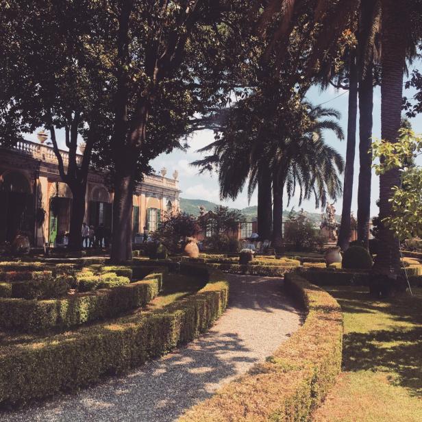 Ein formeller Garten mit geschnittenen Hecken und Palmen vor einem italienischen Herrenhaus.