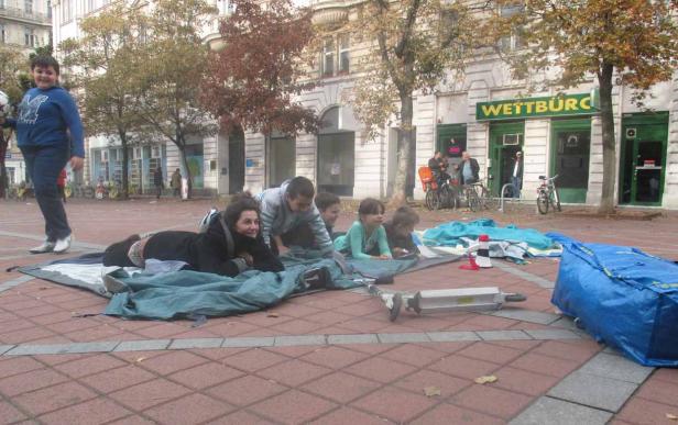 Eine Gruppe von Kindern und eine Frau liegen auf einer Decke auf einem Platz vor einem Wettbüro.